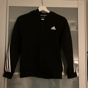Svart Adidas hoodie med vita ränder - Svart hoodie från Adidas med klassiska vita ränder längs ärmarna och över ryggen. Tröjan har dragkedja framtill, huva och Adidas-logga på bröstet. Perfekt för dig som gillar sportig och stilren look.