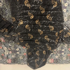 Svart pufferjacka från The North Face - Säljer en svart pufferjacka från The North Face med guldiga loggor över hela jackan. Jackan har huva, dragkedja framtill och två sidofickor med dragkedja. Perfekt för dig som vill sticka ut med en fet och unik vinterjacka.