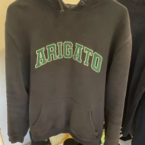 Mörkblå hoodie Axel Arigato - Mörkblå hoodie från Axel Arigato med grön och vit logga framtill. Klassisk känguruficka, huva med snörning och mjukt material. Perfekt för en chill och stilren look.
