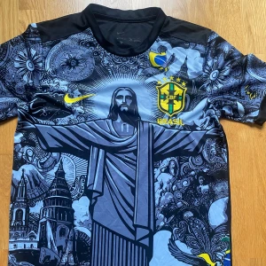 Nike Brasil CBF grafisk fotbollströja - Unik Brasilien CBF fotbollströja från Nike med stor grafisk print av Kristusstatyn och detaljerade illustrationer i svart, grått och blått. Gula broderade loggor och emblem. Kortärmad modell i lätt, ventilerande material. Perfekt för dig som vill sticka ut på planen.