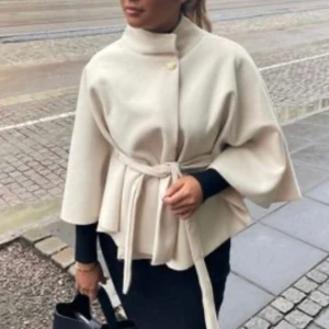 Beige kappa med bälte från Missya - Stilren beige kappa från Missya med hög krage, knäppning med guldig knapp och knytbälte i midjan. Jackan har vida ärmar och en elegant, något utsvängd siluett. Perfekt för dig som vill ha en chic och modern look.