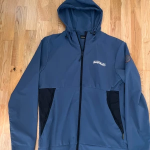 Blå vindjacka från Napapijri med huva - Snygg blå vindjacka från Napapijri med svart detaljer vid fickorna, hel dragkedja och justerbar huva. Jackan har logga på bröstet och patch på ärmen. Perfekt för dig som gillar stilren och sportig look.