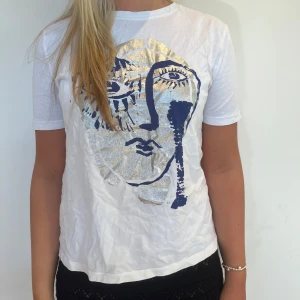 Vit t-shirt med konstnärligt tryck - Vit t-shirt med korta ärmar och ett stort konstnärligt ansikte tryckt i blått och silver på framsidan.