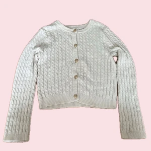 Vit stickad kofta med guldknappar - Supersöt vit stickad kofta med snyggt kabelstickat mönster och guldiga knappar framtill. Koftan har rund halsringning, långa ärmar och ribbade muddar.