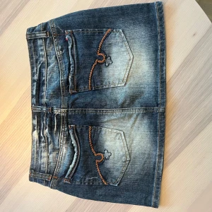 Blå jeanskjol med broderade fickor - Snygg kort jeanskjol i blå denim med slitningar och coola kontrastsömmar. Kjolen har klassiska fem fickor och bakfickor med orange broderi och liten detaljsöm. Perfekt för dig som gillar en avslappnad och trendig stil. Många intresserade så var snabb!!! Kan gå ner i pris vid snabb affär!!