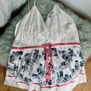 Vit blommigt linne från Du & Jag - Supersött linne med smala axelband och blommigt mönster i mörkblått. Linnet har rosa band och rosett framtill, storlek M.