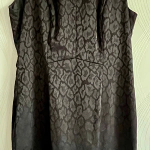 Svart klänning med leopardmönster - Snygg svart klänning med diskret leopardmönster och öppen rygg. Klänningen är ärmlös och har dragkedja bak för enkel påklädning. Materialet har en lätt glansig finish som ger en cool look.