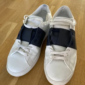Valentino Garavani Open sneakers vit/blå - Snygga Valentino Garavani Open sneakers i vitt skinn med bred marinblå rem över vristen och coola nitar bak på sulan. Klassisk rund tå, vita snören och platt sula. Perfekta för dig som gillar stilrena men ändå unika sneakers. Använda endast en gång. Säljes på grund av att de är för små. Köpta i andrahand. Pris kan förhandlas.
