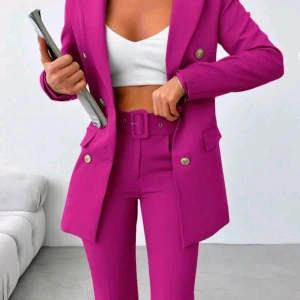 Fuchsia kostym med kavaj och byxa - Säljer en snygg fuchsia kostym med dubbelknäppt kavaj och matchande byxa med bälte. Kavajen har silvriga knappar och fickdetaljer, byxan är högmidjad och smal i modellen. Perfekt för dig som vill sticka ut med färg och stil.