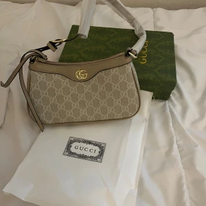 Beige axelväska - Helt ny och oanvänd. Snygg beige axelväska med ett elegant mönster och gulddetaljer. Väskan har en justerbar axelrem och en dekorativ dragkedja. . (Kommer med dustbag och låda) ‼️‼️ pris kan diskuteras‼️‼️