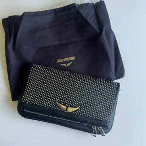 Zadig & Voltaire svart axelväska med nitar - Snygg svart axelväska från Zadig & Voltaire i skinn med silverfärgade nitar och kedja. Väskan har dubbla dragkedjor, ikonisk vingedetalj i metall och medföljer dustbag. Perfekt för dig som gillar edgy och trendig stil.