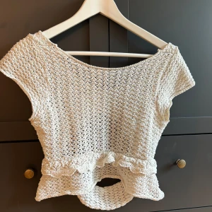 Vit stickad topp med volang från Zara - Supersöt vit stickad topp från Zara med korta ärmar och volangdetalj i midjan. Toppen har ett öppet parti med knytning baktill och är gjord i ett luftigt, virkat material. Perfekt för dig som gillar unika detaljer och en somrig vibe!
