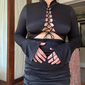 Svart kort klänning med snörning - Svart kort klänning med lång ärm och utsvängda ärmslut. Djup snörning framtill och cut-out under bysten ger en edgy vibe. Sitter tight och har en lätt glansig finish, perfekt för dig som vill sticka ut.
