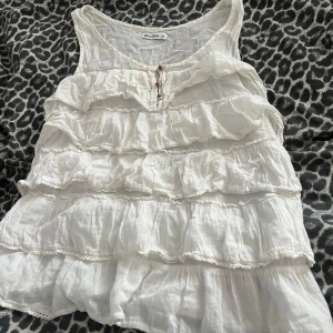 Vit volanglinne från miss Milla  - Supersött vitt linne med flera volanglager och knappar framtill från miss Milla . Luftigt material och perfekt för varma dagar. Rund halsringning och ärmlös design ger en avslappnad och trendig look.