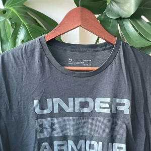 Under Armour t-shirt - Svart t-shirt från Under Armour med stor logga och text framtill i grått. Klassisk rund halsringning och korta ärmar. Tillverkad i mjuk bomull, perfekt för träning eller chill. Snygg och enkel stil som funkar till det mesta.