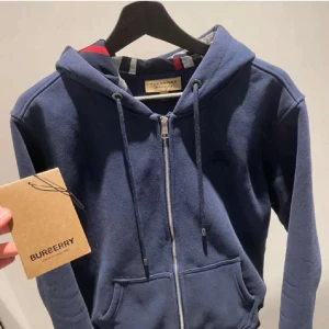 Blå hoodie med dragkedja från Burberry - Snygg marinblå hoodie från Burberry med hel dragkedja, snörning i huvan och klassiskt rutigt foder på insidan. Tröjan har känguruficka framtill och en relaxed passform. Perfekt för dig som gillar stilrena och exklusiva streetwear-plagg.