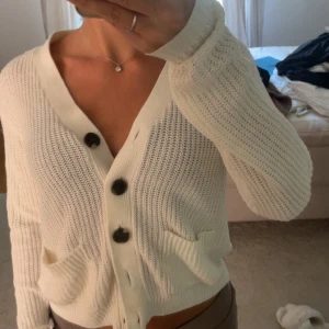 Vit stickad kofta med stora knappar - Superfin vit stickad kofta med stora svarta knappar och två fickor framtill. Koftan har v-ringning, lång ärm och en lite croppad passform som känns både chill och trendig. Perfekt att slänga på sig över ett linne eller t-shirt.