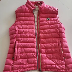 Rosa dunväst från North Sails - Snygg rosa dunväst från North Sails med quiltad design och hög krage. Västen har dragkedja framtill, två fickor och en liten logotyp på bröstet. Perfekt att slänga över en hoodie för en sportig look.