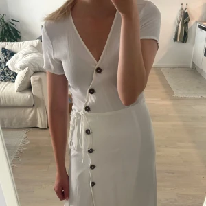 Vit omlottklänning från Bik Bok - Superfin vit omlottklänning från Bik Bok! Den har knappar i fram och band så att man kan knyta en liten rosett på sidan och därmed justera hur den ska sitta runt midjan.   Storlek XS men passar mig som är S också.