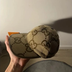 Beige keps från Gucci med GG-mönster - Snygg beige keps från Gucci med det klassiska GG-monogrammet i brunt över hela kepsen. Kepsen har böjd skärm och är tillverkad i ett tåligt tygmaterial. Perfekt accessoar för att lyfta din outfit.