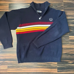 Randig stickad tröja Fred Perry - Mörkblå stickad tröja från Fred Perry med half zip och högt krage. Snygga ränder i rött, gult och vitt över bröstet och ryggen. Lite retro och perfekt nu till hösten. 