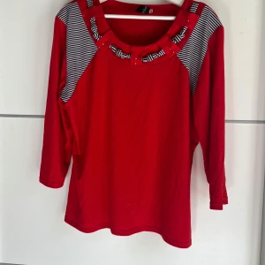 Röd långärmad tröja från Roma - Röd långärmad tröja från Roma med svartvita ränder på axlarna och runt den runda halsringningen. Snygga pärldetaljer på kragen ger extra edge. Skön passform och tillverkad i mjukt bomullsmaterial. Perfekt för dig som gillar färg och unika detaljer.