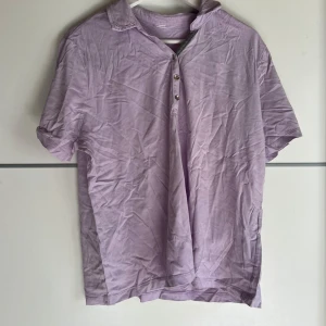 Ljuslila pikétröja med knappar - Ljuslila pikétröja med klassisk krage och tre knappar framtill. Tröjan har korta ärmar med knappdetalj och är gjord i mjukt bomullstyg. Perfekt för dig som gillar en chill och stilren look.