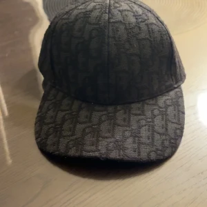 Svart keps med mönster - Snygg svart keps med diskret mönster i tyget som ger en exklusiv känsla. Kepsen har klassisk form med böjd skärm och är tillverkad i ett grovt tyg som påminner om bomull eller polyester. Perfekt accessoar för att lyfta din outfit.