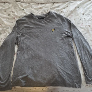 Grå långärmad tröja Lyle & Scott - Snygg grå långärmad tröja från Lyle & Scott med klassisk rund hals och ikonisk gul logga på bröstet. Tillverkad i mjuk bomull som är skön mot huden. Perfekt basic-plagg med normal passform som funkar året runt.