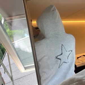 Grå hoodie med svart tryck och stjärna - Snygg ljusgrå hoodie med svart texten 'AMOUR' framtill och en cool svart stjärna på ryggen. Tröjan har huva med snörning och är långärmad. Perfekt för chill dagar och enkel att matcha med jeans eller mjukisbyxor.