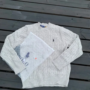 Grå stickad tröja får Ralph Lauren  - Exklusiv kabelstickad tröja från Polo Ralph Lauren – grå Stilren lgrå stickad tröja i klassisk kabelmönster från Polo Ralph Lauren. Tillverkad i mjuk bomull med den ikoniska mörkblå Polo-loggan broderad på bröstet. Tröjan har en rund halsringning och ribbade muddar för en tidlös och bekväm passform. Ny med tag – perfekt för både vardag och finare tillfällen.
