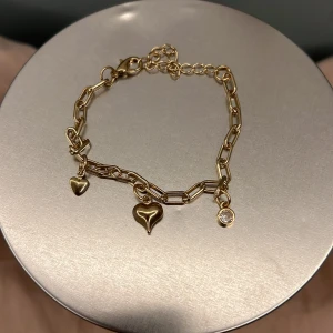  Guldigt armband med hjärtberlocker - Snyggt armband i guldton med chunky kedja och tre berlocker: två hjärtan och en liten glittrig sten. Perfekt accessoar för att lyfta din outfit med en söt och trendig touch.