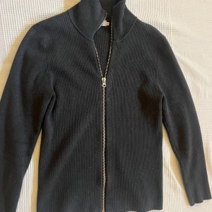 Fullzip kofta från Ljung mörkgrå - Mörkgrå ribbad kofta från Ljung by Marcus Larsson i storlek S. Koftan har hel dragkedja framtill, hög krage och är långärmad. Materialet är mjukt och stretchigt.