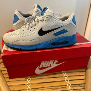 Nike Air Max 90 Premium Comfort blå - Helt nya Nike Air Max 90 Premium Comfort sneakers i vitt och blått med svarta detaljer. Ovandel i mesh och syntet, klassisk Air Max-dämpning och svart yttersula med blå inslag. Snygg sportig look med tydlig Nike-logga på häl och plös. Perfekta för dig som gillar streetstyle.UTGÅENDE MODELL