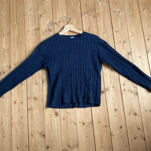 Mörkblå kabelstickad tröja Lindex - Kabelstickad tröja från Lindex i en snygg mörkblå färg. Tröjan har rund halsringning, ribbade muddar och lång ärm. Perfekt för dig som gillar klassisk stil med en twist. Materialet är mjukt och skönt, passar till många outfits.