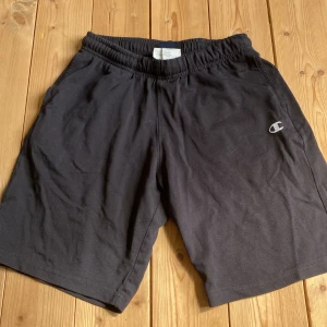 Svarta Champion shorts i bomull - Svarta shorts från Champion med elastisk midja och diskret logga på ena benet. Tillverkade i mjuk bomull, perfekta för chill dagar eller träning. Klassisk passform och enkel stil som funkar till det mesta.