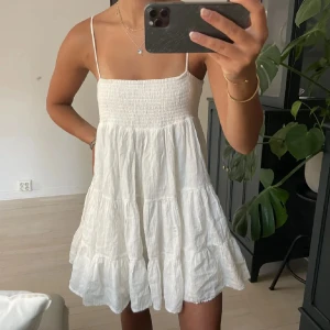 Vit somrig klänning från Zara - Supersöt vit kortklänning från Zara med smock upptill och tunna axelband. Klänningen har volangig kjol och är gjord i ett lätt, luftigt material som passar perfekt för varma dagar. Enkel och stilren design som känns fräsch och trendig.