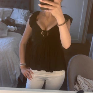 Svart blus med knyt och volang - Supersöt svart blus med djup v-ringning och knyt framtill. Toppen har volangiga axelband och ett flowigt fall nedtill som ger en romantisk vibe. Perfekt för dig som gillar feminina detaljer och vill sticka ut lite extra.