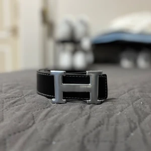 Svart Bälte med H-spänne - Stilrent svart bälte med H-spänne i silverfärgad metall. Bältet är gjort i skinn med vit kontrastsöm längs kanterna och har en klassisk, tidlös design som lyfter vilken outfit som helst.