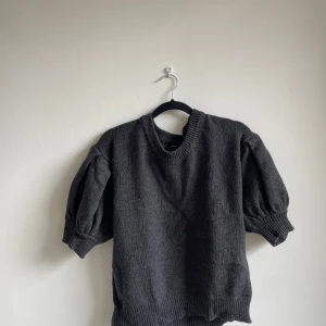 Mörkgrå stickad tröja med puffärm Lindex - Mörkgrå stickad tröja från Lindex med korta puffärmar och bred ribbad kant vid ärmslut och nederkant. Tröjan har rund halsringning och en loose fit som ger en chill vibe. Perfekt att styla med jeans eller kjol för en trendig look.