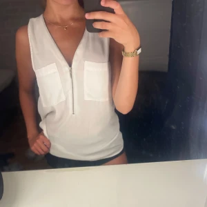 Vit ärmlös linnetopp med dragkedja - Snygg vit ärmlös linnetopp med två stora bröstfickor och en djup v-ringning med dragkedja framtill. Perfekt till sommaren och enkel att matcha med både jeans och shorts. Lätt och luftig känsla. Skriv för fler frågor!