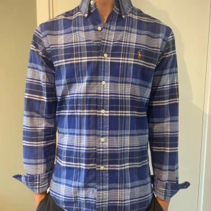 Blårutig skjorta från Ralph Lauren - Snygg blårutig skjorta från Ralph Lauren i slim fit-modell. Skjortan har klassisk krage, långa ärmar och knappar framtill. Ikoniska broderade loggan på bröstet. Perfekt för dig som gillar stilrena och tidlösa plagg.