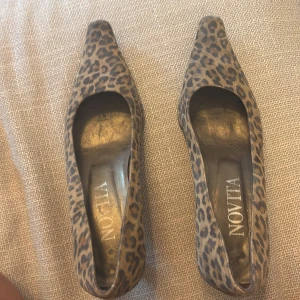 Leopardmönstrade pumps från Novita - Snygga pumps från Novita i leopardmönstrad mocka med spetsig tå och låg klack. Skorna har en brun och svart färgskala och ger en cool statement-look. Perfekta för dig som vill sticka ut med djurmönster och elegant form.