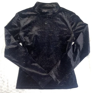 Glittrig svart polotröja från Monki - Svart polotröja från Monki i glittrigt sammetstyg. Tröjan har lång ärm och en hög krage som ger en snygg siluett. Perfekt för dig som vill sticka ut med lite extra glitter. Materialet är mjukt och stretchigt för en bekväm passform.