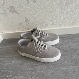 Grå mockasneakers från Axel Arigato - Snygga sneakers från Axel Arigato i ljusgrå mocka med vita, platta sulor och klassisk snörning. Minimalistisk design med rund tå och diskret logga i guld på sidan. Perfekta för en clean och stilren look. Burits en gång innan!