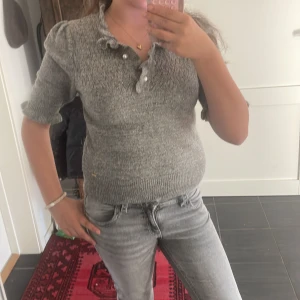 Grå stickad kortärmad tröja med volang - Superfin grå stickad tröja med korta ärmar och volangdetaljer vid halsringningen och ärmsluten. Tröjan har pärlknappar framtill och en feminin, figurnära passform. Perfekt för dig som gillar en söt och stilren look.