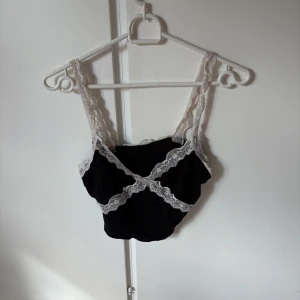 Svart croppad linnetopp med spets - Supersöt svart croppad linnetopp med vita spetsdetaljer längs axelbanden och runt kanterna. Toppen har en figurnära passform och är perfekt för att skapa en trendig och feminin look. Spetsen ger en romantisk vibe och toppen är ärmlös med breda axelband.
