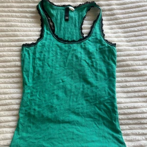 Grön linnetopp med spetskant - Snygg grön linnetopp med svarta spetsdetaljer längs kanterna. Modellen är ärmlös med brottarrygg och har en lätt ribbad struktur i tyget. Perfekt för dig som gillar en enkel men ändå detaljrik look.