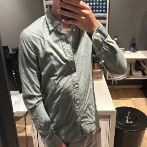 Grå skjorta från Tommy Hilfiger - Snygg grå skjorta från Tommy Hilfiger med klassisk krage och vita knappar. Skjortan har lång ärm och en liten broderad logga på bröstet. Tillverkad i mjuk bomull, perfekt för en clean och stilren look.