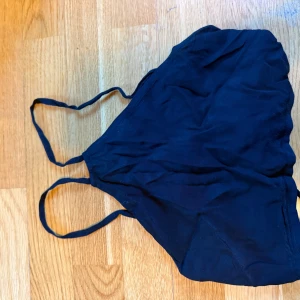 Svart omlott-linnetopp från Gina Tricot - Svart croppad linne-topp från Gina Tricot med tunna axelband och snygg omlottknytning i ryggen. Toppen har en halterneck-liknande skärning framtill och smala band som korsas och knyts baktill. Perfekt för sommarens varma dagar. På bilderna har jag ett linne under! Man kan ändra knytningen i ryggen hur man vill! Använt en gång. 